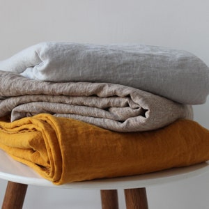 May include: A stack of three linen throws in different colours: mustard yellow, beige, and light grey. The throws are folded and stacked on a white stool.