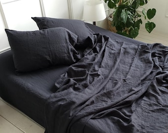 May include: A black linen bedding set with a flat sheet, fitted sheet, and two pillowcases. The bedding is arranged on a bed with a white headboard and white wooden floor.