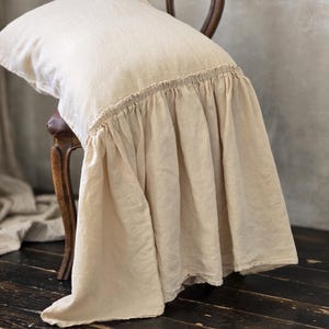 May include: A cream-colored linen pillow with a ruffled skirt detail, resting on a vintage wooden chair. The pillow has a soft, textured appearance, and the chair has a curved back.