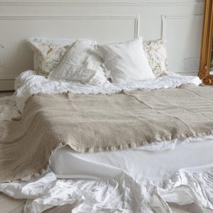 May include: A bed with white linens, pillows, and a beige knitted throw. The pillows have floral and solid white patterns. The bed skirt has a ruffled design. The room has white walls and a wooden floor.