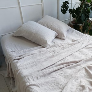 May include: A bed with light beige linen bedding, including a fitted sheet, flat sheet, and two matching pillowcases. The sheets have a slightly wrinkled texture, suggesting a natural fabric. The bed is in a bright room with a white headboard.