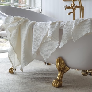 Puede incluir: Una bañera blanca con patas de garra doradas y grifería dorada. Una toalla blanca con tejido de gofre cuelga del borde. Un pequeño jarrón con flores blancas completa la escena, creando un ambiente relajante.