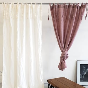 May include: Two linen curtains hanging from a rod. The curtain on the left is white and the curtain on the right is a dusty rose color. Both curtains have a gathered look with ties at the top.
