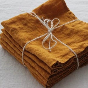 Puede incluir: Una pila de seis servilletas de lino amarillo dorado dobladas, atadas con un lazo de cuerda blanca. Las servilletas están cuidadosamente dispuestas, mostrando su superficie texturizada. El fondo es una tela blanca lisa.
