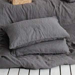 May include: Dark grey linen bedding set, including a duvet cover and two pillowcases. The bedclothes are arranged on a white wooden surface. A small woven basket is visible in the background.