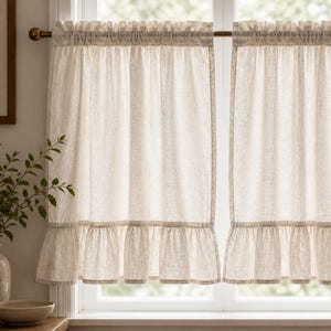 Peut inclure: Deux rideaux de cuisine crème à volants suspendus à une fenêtre. Les rideaux ont un design simple de style champêtre avec un bord inférieur à volants et une bordure décorative. Les rideaux sont en tissu de couleur claire.