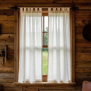 Op de afbeelding: Doorzichtige witte linnen gordijnen hangen in een houten raamkozijn. De gordijnen laten natuurlijk licht door. Het raam staat in een rustieke, houten wand, wat een gezellige, boerderijachtige sfeer creëert.