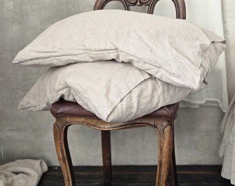 May include: Two beige linen pillows stacked on a wooden chair with a brown leather seat. The chair has a carved design and sits on a dark wood floor.