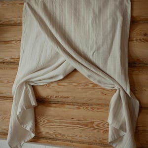 May include: A light beige, semi-sheer curtain panel with a subtle vertical striped pattern. The fabric is draped over a horizontal rod, creating a crossed, overlapping effect in the center that leaves the lower corners hanging down loosely.