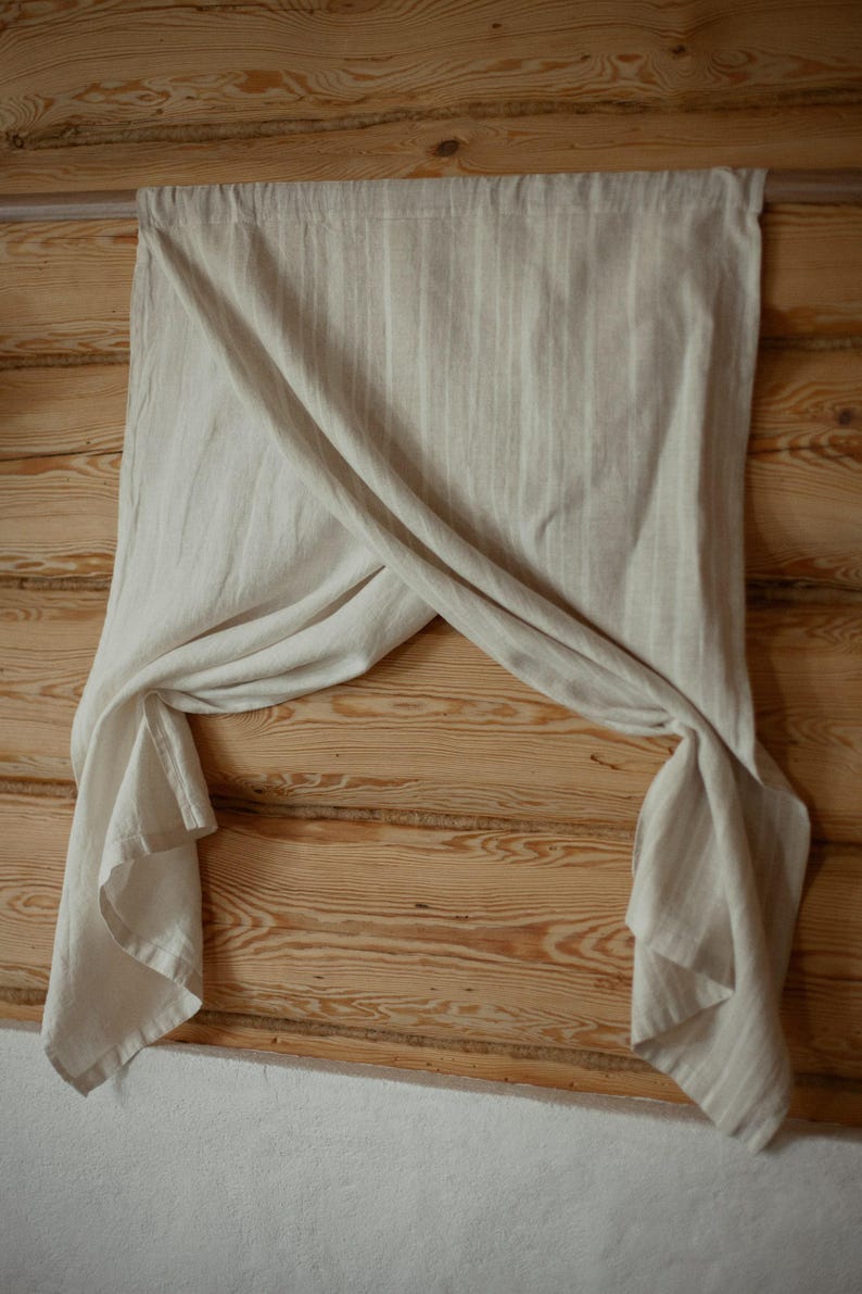 May include: A light beige, semi-sheer curtain panel with a subtle vertical striped pattern. The fabric is draped over a horizontal rod, creating a crossed, overlapping effect in the center that leaves the lower corners hanging down loosely.