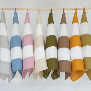 May include: A collection of eight striped kitchen towels in various colors, including gray, blue, pink, beige, olive green, brown, orange, and yellow. Each towel is hanging from a wooden rod, showcasing a clean and organized display.