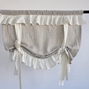 Könnte beinhalten: Ein Fensterbehang aus Leinen in neutralen Tönen. Er hat einen grauen oberen Bereich und einen helleren Hauptteil. Weiße Rüschen und Bindebänder sorgen für Dekoration. Der Behang hängt an einer schwarzen Stange.