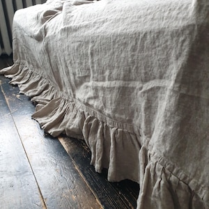 May include: A beige linen sofa throw with a ruffled edge. The throw is draped over a sofa and the ruffled edge is visible on the floor.