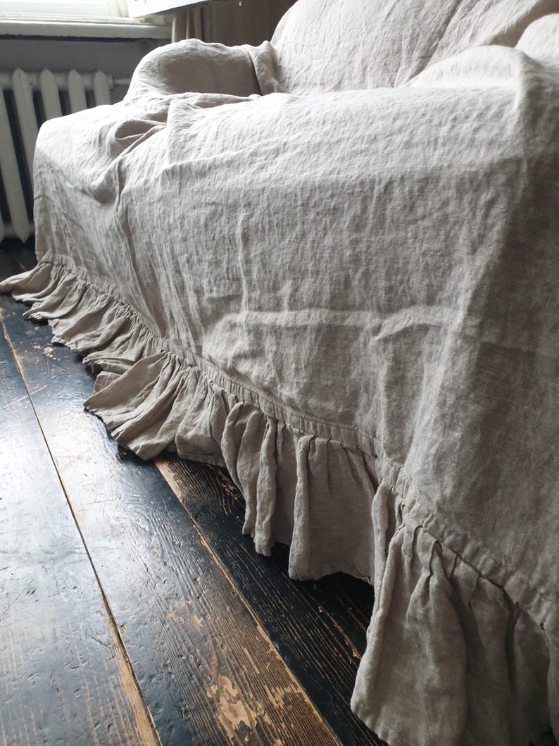 May include: A beige linen sofa throw with a ruffled edge. The throw is draped over a sofa and the ruffled edge is visible on the floor.