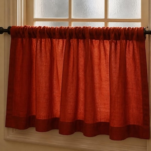 May include: A pair of rust-colored window curtains. The curtains are made of a textured fabric and are hung on a dark rod. The bottom of the curtains have a darker band of fabric. The curtains are in front of a window with frosted glass.