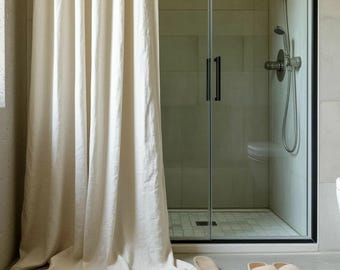 May include: A bathroom scene featuring a cream-colored shower curtain and a glass shower enclosure. Two pairs of beige slippers rest on the gray floor. The showerhead and fixtures are visible inside the enclosure.