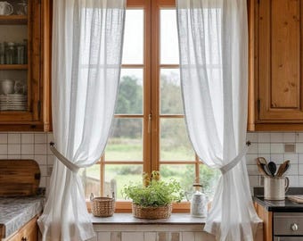 May include: A kitchen window with sheer white curtains tied back with fabric ties. The wooden window frame and surrounding cabinetry are a warm, natural wood tone. A small basket and potted plant sit on the windowsill, adding a touch of greenery.