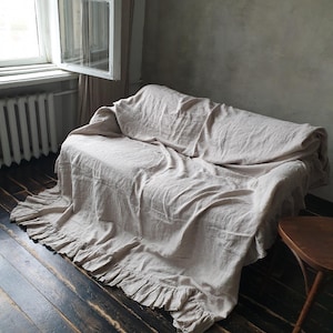 May include: A light beige linen throw blanket with a ruffled edge, draped over a couch in a room with a window and wooden floor.