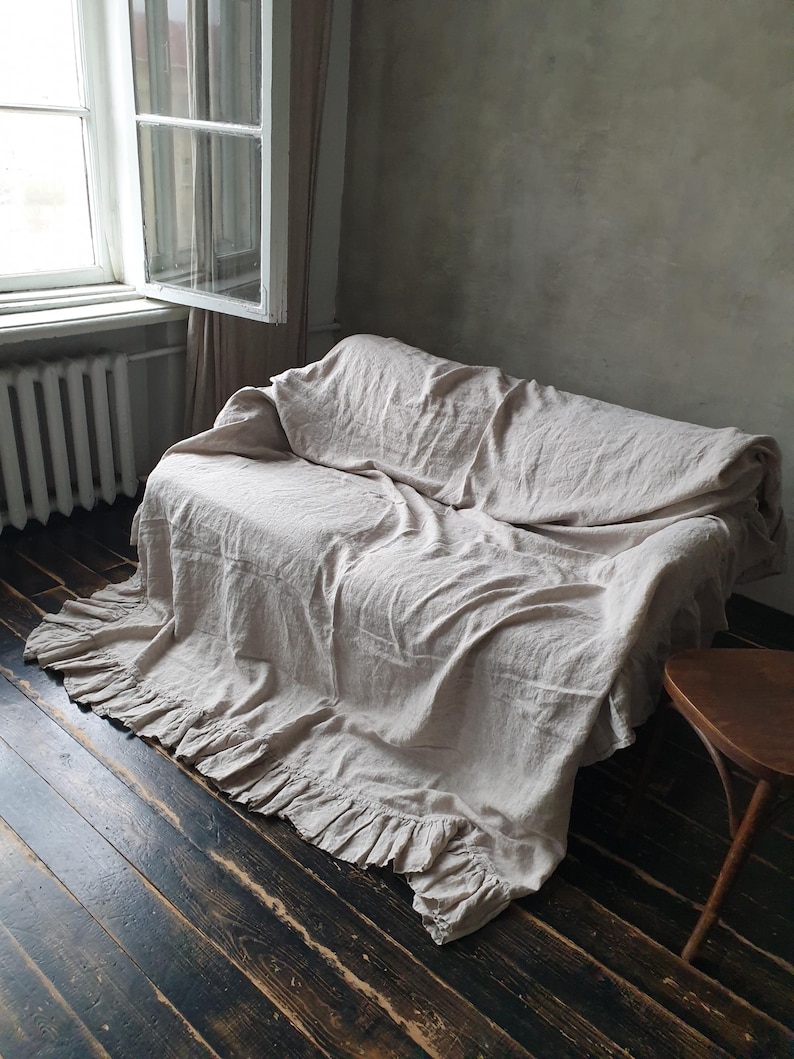 May include: A light beige linen throw blanket with a ruffled edge, draped over a couch in a room with a window and wooden floor.