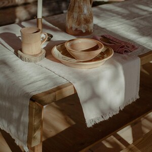 May include: A rustic wooden table setting with a white linen tablecloth and a brown ceramic pitcher, bowl, and mug. A pink napkin is placed next to the bowl.