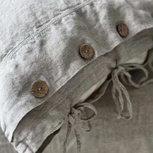 Peut inclure: Gros plan sur une taie d'oreiller en lin gris clair avec trois boutons en bois et des attaches à nouer. La taie d'oreiller a un aspect texturé et un design simple et minimaliste, adapté à la décoration de la chambre.