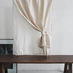May include: A rectangular curtain panel made of off-white fabric with vertical beige stripes. The curtain is suspended from a silver metal rod and is gathered to one side with a matching fabric tie, creating a draped effect across the center.