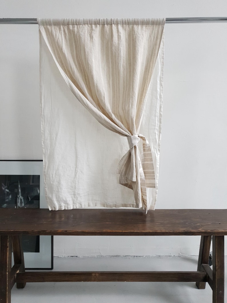 May include: A rectangular curtain panel made of off-white fabric with vertical beige stripes. The curtain is suspended from a silver metal rod and is gathered to one side with a matching fabric tie, creating a draped effect across the center.