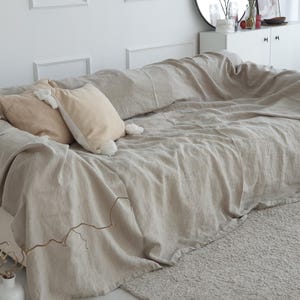 Può includere: Una coperta in lino beige chiaro drappeggiata su un divano con due cuscini beige. La coperta ha un aspetto testurizzato e intrecciato ed è piegata sullo schienale del divano.