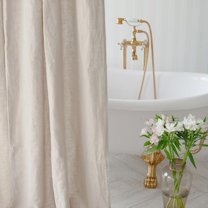 May include: A white bathtub with gold fixtures and a beige linen shower curtain. White flowers in a clear glass vase are on the floor next to the tub.