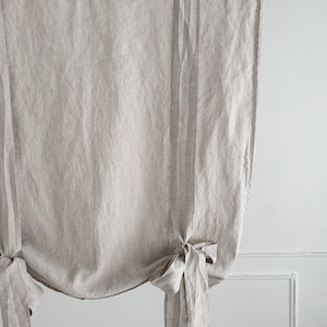 May include: A pair of beige linen roman shades with a gathered, tied-back design. The shades are hung on a black rod.