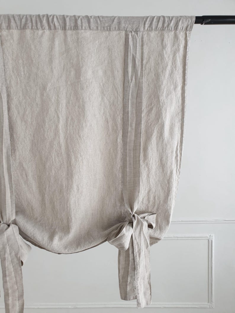 May include: A pair of beige linen roman shades with a gathered, tied-back design. The shades are hung on a black rod.