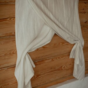 May include: A light beige curtain featuring a subtle vertical striped pattern. The fabric is draped and gathered, creating soft folds that hang from a rod. The material has a textured appearance with a neutral color palette throughout the piece.