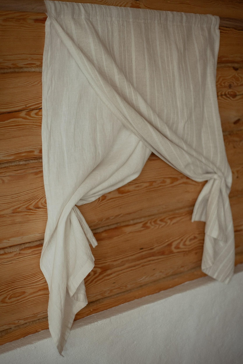 May include: A light beige curtain featuring a subtle vertical striped pattern. The fabric is draped and gathered, creating soft folds that hang from a rod. The material has a textured appearance with a neutral color palette throughout the piece.