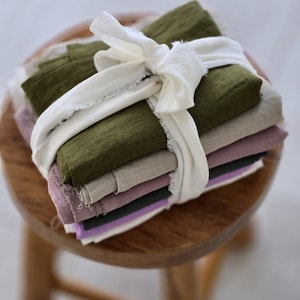 May include: A stack of folded fabric in various colors, including olive green, lavender, and beige, tied with a white ribbon. The fabric is arranged on a small wooden stool, creating a visually appealing display of textiles.