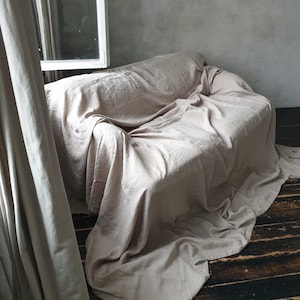 May include: A light beige linen sofa throw blanket draped over a couch, with the edges of the blanket hanging down to the floor. The couch is in a room with a window and a curtain.