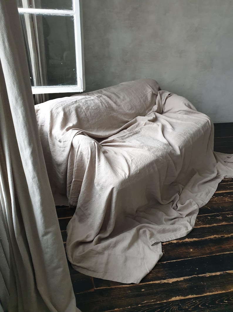May include: A light beige linen sofa throw blanket draped over a couch, with the edges of the blanket hanging down to the floor. The couch is in a room with a window and a curtain.