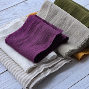 Könnte beinhalten: Eine Sammlung gefalteter Leinenstoffe in verschiedenen Farben. Die Stoffe umfassen Farbtöne von Lila, Beige, Olivgrün und Senfgelb. Einige Stoffe haben einen rohen Rand, und einer hat ein gestreiftes Muster. Die Stoffe sind auf einer hellblauen Holzoberfläche gestapelt.