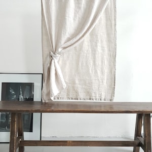 May include: A rectangular curtain featuring a vertical beige and white striped pattern. The fabric is gathered and tied into a knot on the left side, creating a draped effect. The curtain hangs from a horizontal metal rod against a plain wall.