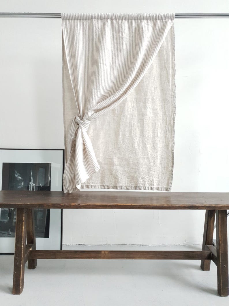 May include: A rectangular curtain featuring a vertical beige and white striped pattern. The fabric is gathered and tied into a knot on the left side, creating a draped effect. The curtain hangs from a horizontal metal rod against a plain wall.