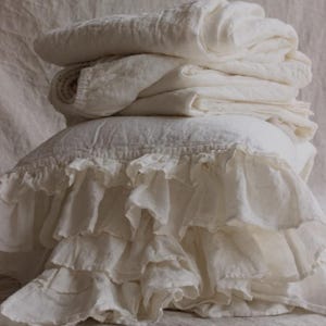 May include: A stack of ivory-colored bedding, including folded sheets and a pillow with ruffled trim. The linens appear to be made of a soft, textured fabric, creating a cozy and inviting aesthetic. The neutral color palette suggests a focus on comfort and simplicity.