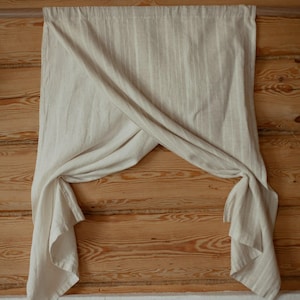 May include: A light beige curtain featuring a subtle vertical striped pattern. The fabric is draped over a horizontal rod, with the two lower corners gathered and tied to create a crossed, overlapping effect in the center of the panel.