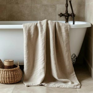 以下が含まれることがあります： 白いバスタブの縁にかけられたベージュのリネンタオル。タオルは質感があり、タイルの床に向かって垂れ下がっています。籐のバスケットと小さな容器が見えます。