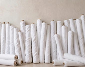 May include: A collection of numerous white fabric bolts arranged in rows, with a wooden yardstick standing vertically on the left side. The fabric rolls are cylindrical, featuring a textured white surface and cardboard cores at the ends.