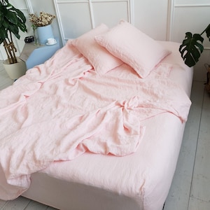 May include: A bed dressed with light pink linen bedding, including a fitted sheet, two pillowcases, and a top sheet. The bedding appears slightly creased. The room has white walls and a wooden floor.