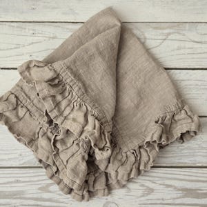 Peut inclure: Une serviette en lin beige pliée avec des bords à volants. La serviette est faite d'un tissu doux et texturé et est placée sur une surface en bois blanc. Les volants ajoutent un élément décoratif.