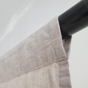 May include: Close-up of a beige linen curtain with a black metal rod. The curtain is hanging from the rod and has a folded edge.