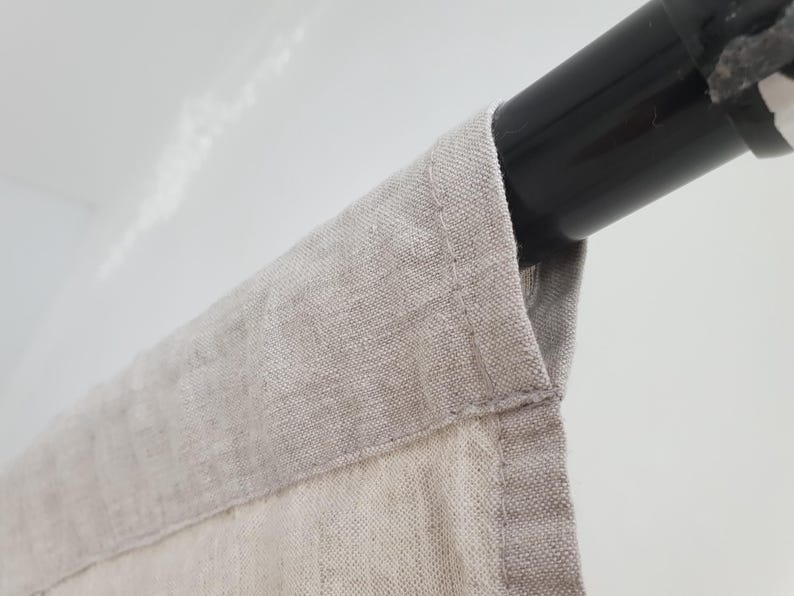 May include: Close-up of a beige linen curtain with a black metal rod. The curtain is hanging from the rod and has a folded edge.