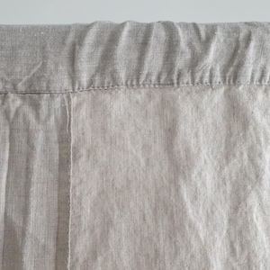 May include: A close-up of a light brown linen curtain with a seam running down the center. The curtain has a folded top edge.