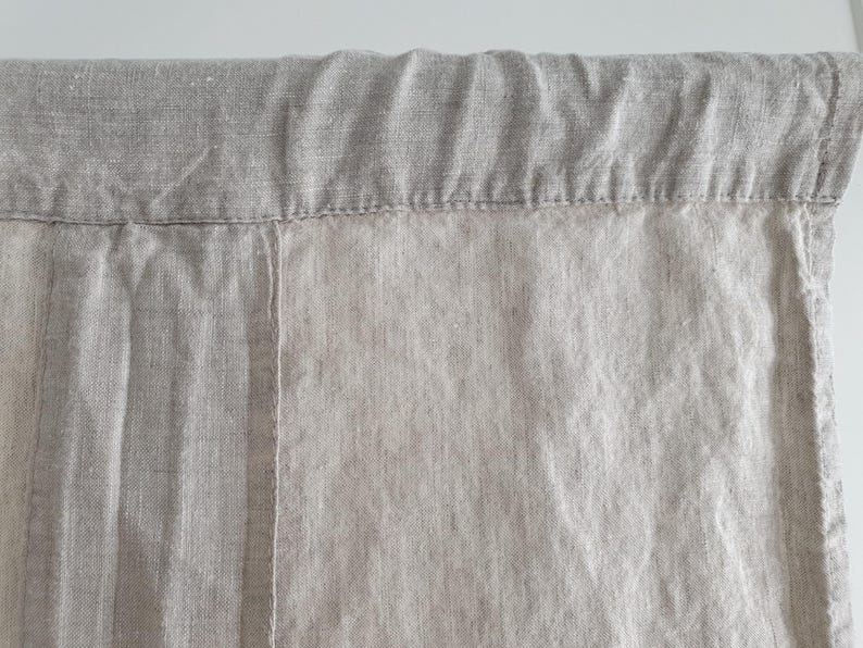 May include: A close-up of a light brown linen curtain with a seam running down the center. The curtain has a folded top edge.