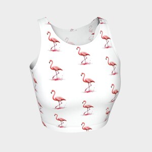 May include: White crop top featuring a repeating pattern of pink flamingos. Each flamingo is depicted in a watercolor style, with a pink body and long legs. The design includes pink splashes around the flamingos, creating a playful and summery aesthetic.